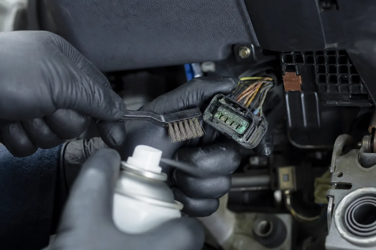 Close-up of technician cleaning corroded electrical connector pins under a vehicle dashboard, 1200x800